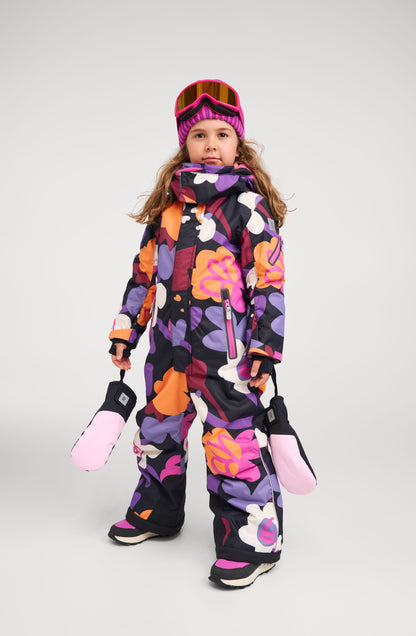 Full-body lifestyle shot of a young girl wearing the Reima Reach kids' ski suit in a vibrant black, purple, orange, and pink floral print. She is geared up for the slopes, holding pink winter mittens and wearing ski goggles, demonstrating the suit's comfortable fit and ease of movement.
