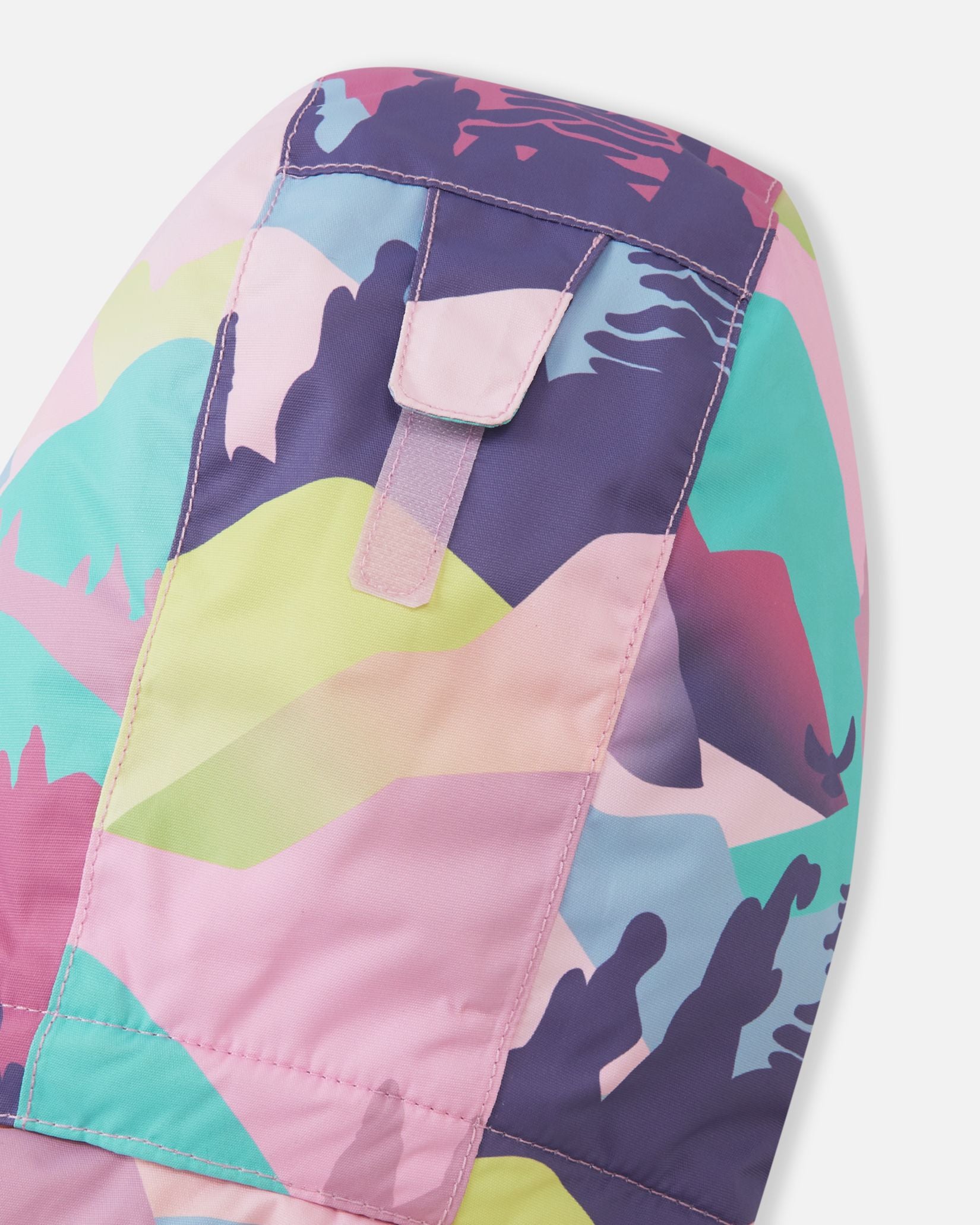 A detailed close-up view of the technical fabric on the hood of a Reima Reach kids' ski suit, featuring an abstract, colorful camouflage-style mountain print in shades of grey, pink, purple, and teal. Stitching and a protective closure flap with snap buttons are visible.