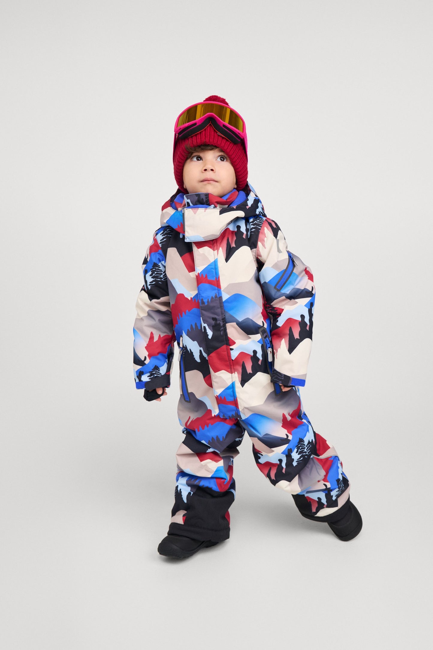 Full-body shot of a young child modeling the Reima Reach ski suit in the soft black, red, and blue mountain print. The child is styled with a red beanie and ski goggles, demonstrating the suit's comfortable fit, mobility, and how easily it accommodates winter accessories for a day on the slopes.