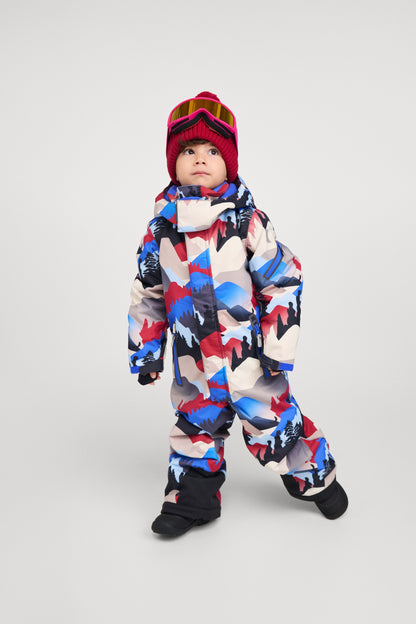 Full-body shot of a young child modeling the Reima Reach ski suit in the soft black, red, and blue mountain print. The child is styled with a red beanie and ski goggles, demonstrating the suit's comfortable fit, mobility, and how easily it accommodates winter accessories for a day on the slopes.