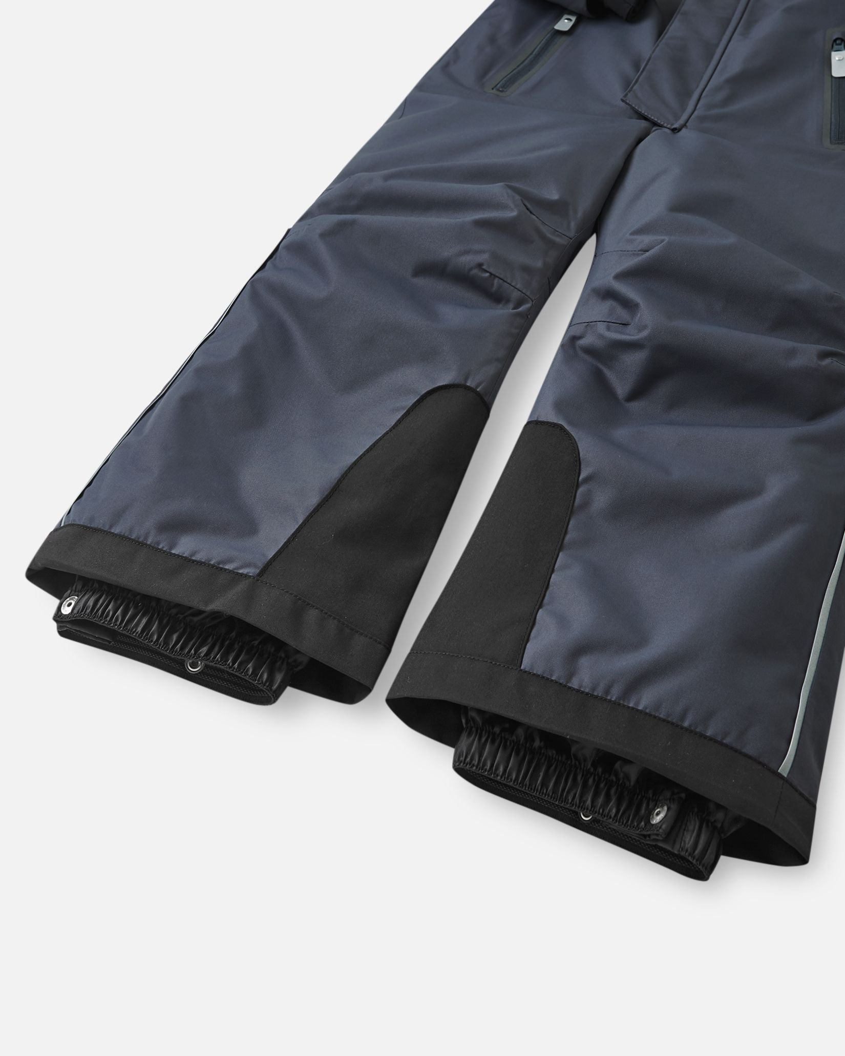 Close-up of the lower legs on the Reima Reach kids' winter ski suit in soft black, showcasing heavy-duty black reinforced kick panels and elastic inner snow gaiters designed to seal tightly over ski boots and keep powder out.