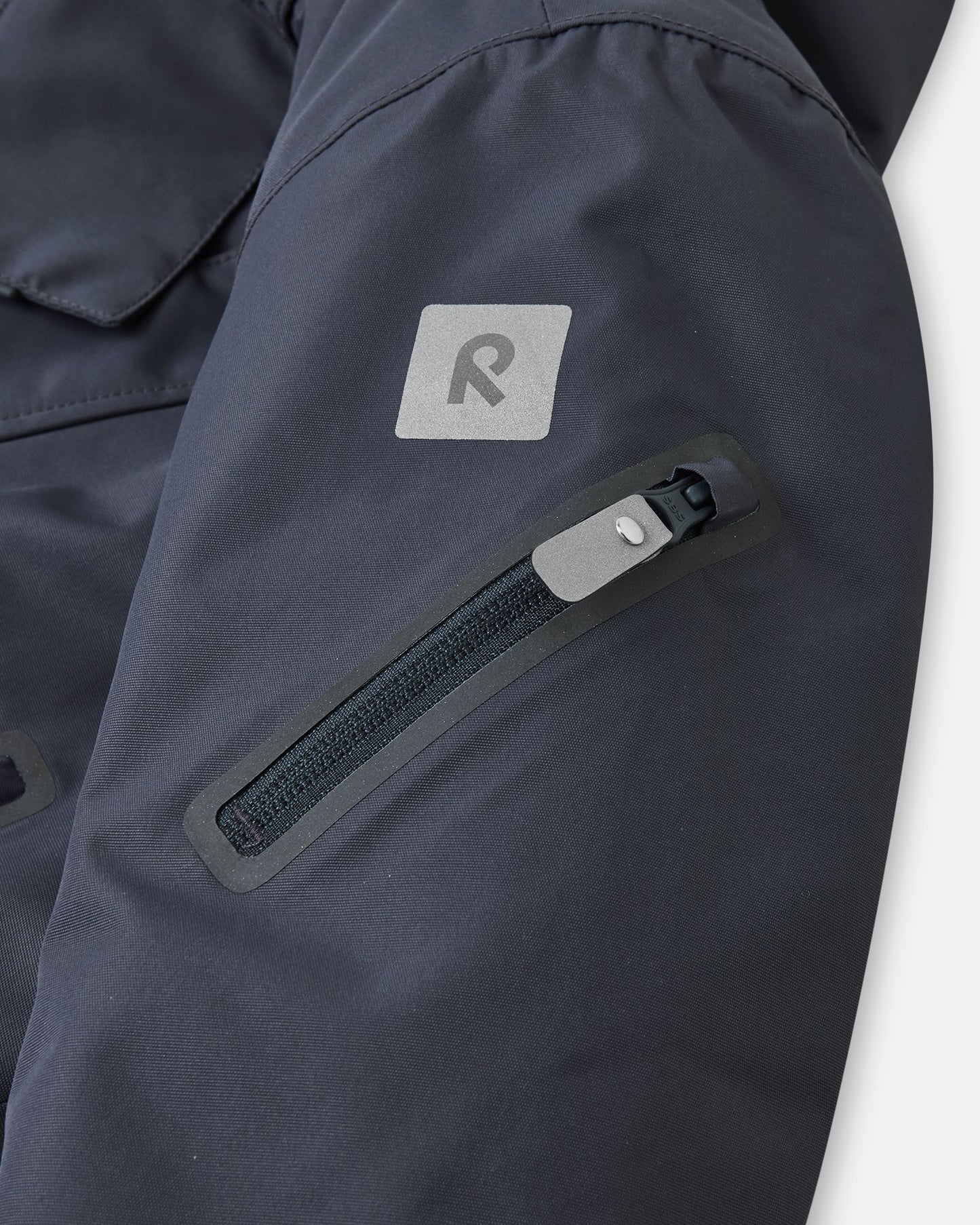 Detail shot of the sleeve on the soft black Reima Reach kids' ski suit, featuring a secure zippered ski pass pocket on the forearm and a high-visibility reflective Reima safety patch for dark winter days.