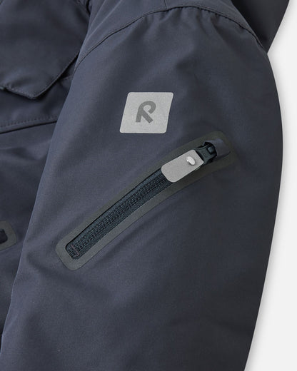 Detail shot of the sleeve on the soft black Reima Reach kids' ski suit, featuring a secure zippered ski pass pocket on the forearm and a high-visibility reflective Reima safety patch for dark winter days.