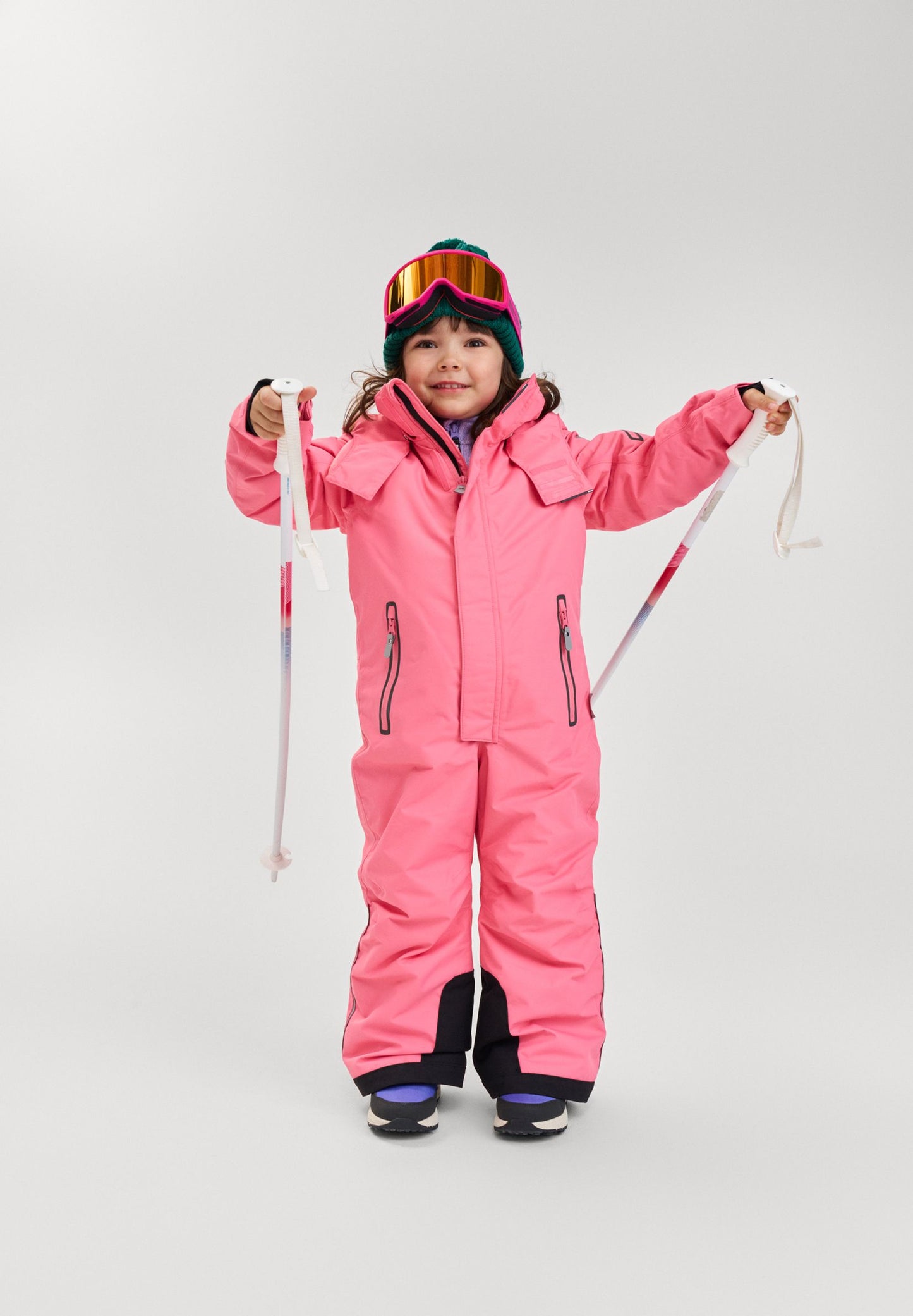 Young girl holding ski poles and wearing the soft coral Reima Reach waterproof ski suit, demonstrating its flexible, ergonomic fit designed specifically for active alpine skiing and winter sports.
