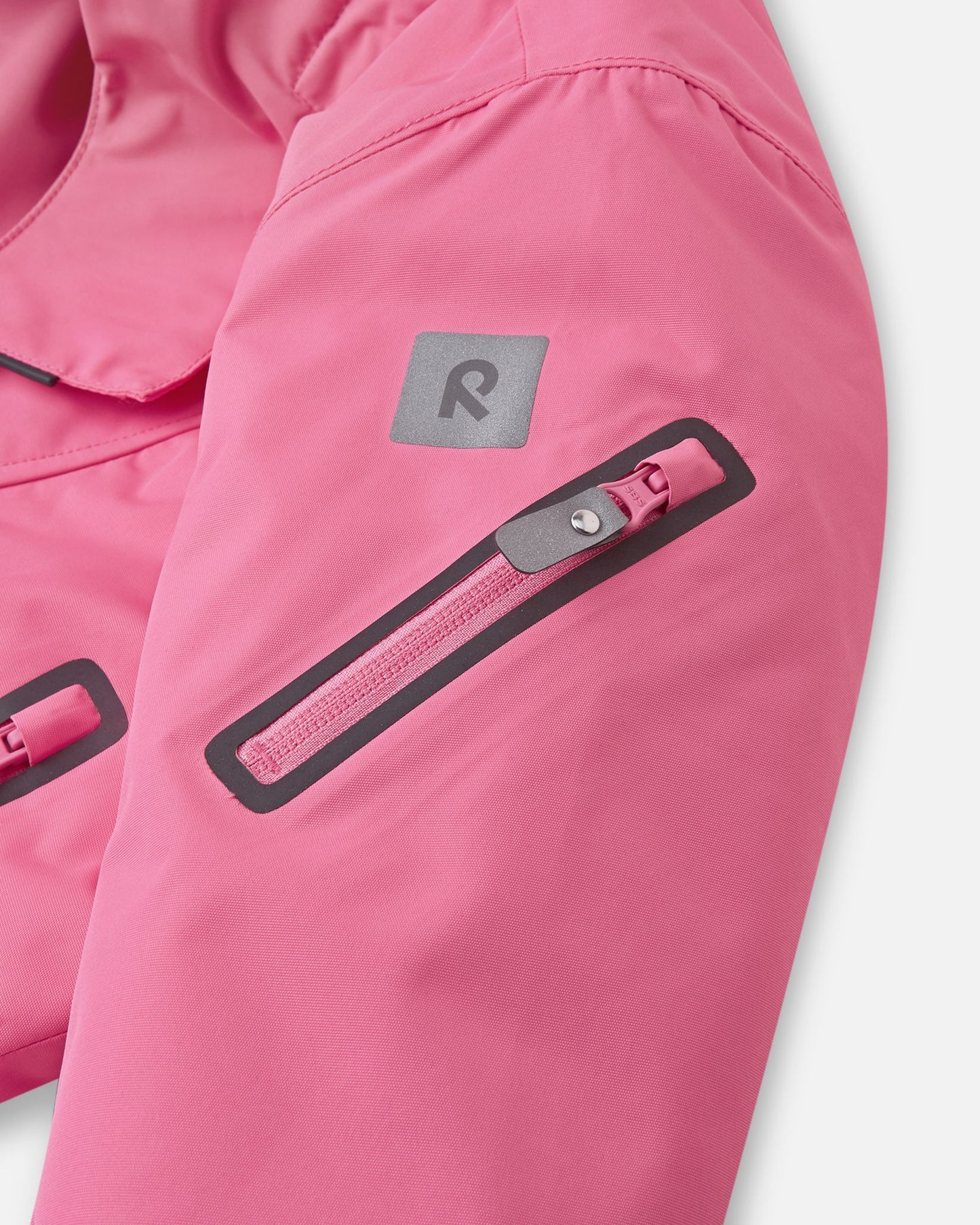 Close-up of the sleeve on the soft coral Reima Reach ski suit, featuring a secure zippered ski pass pocket on the forearm and a high-visibility reflective Reima logo patch.
