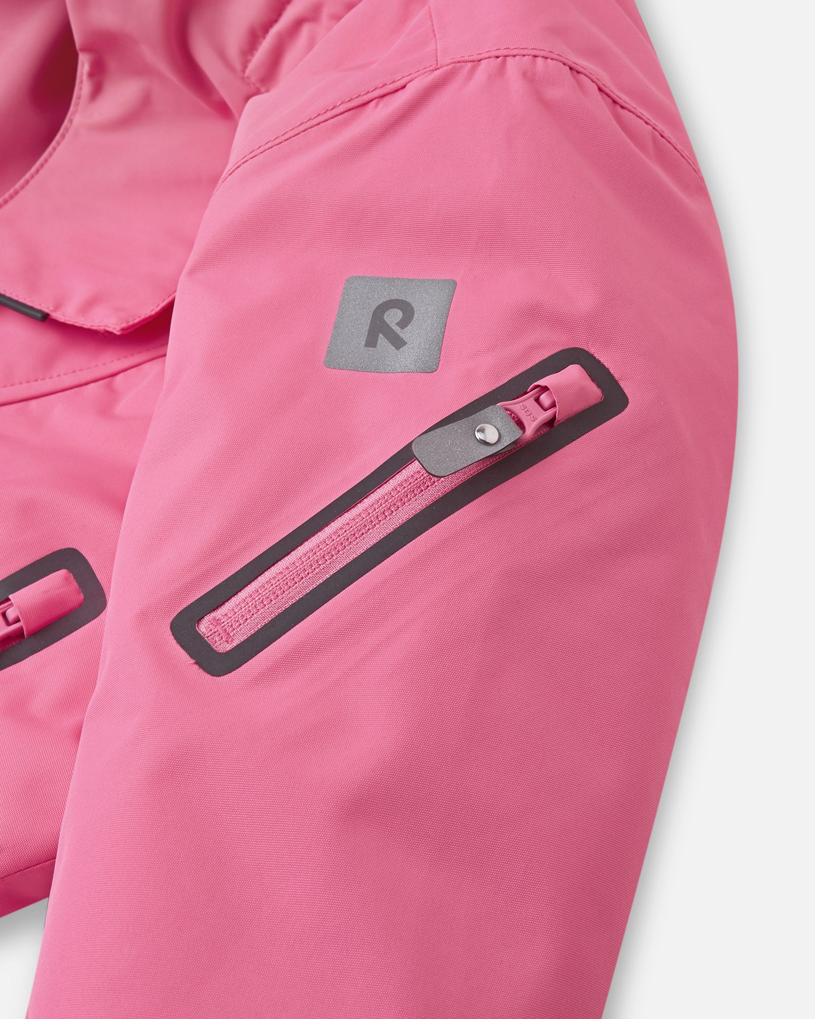 Close-up of the sleeve on the soft coral Reima Reach ski suit, featuring a secure zippered ski pass pocket on the forearm and a high-visibility reflective Reima logo patch.