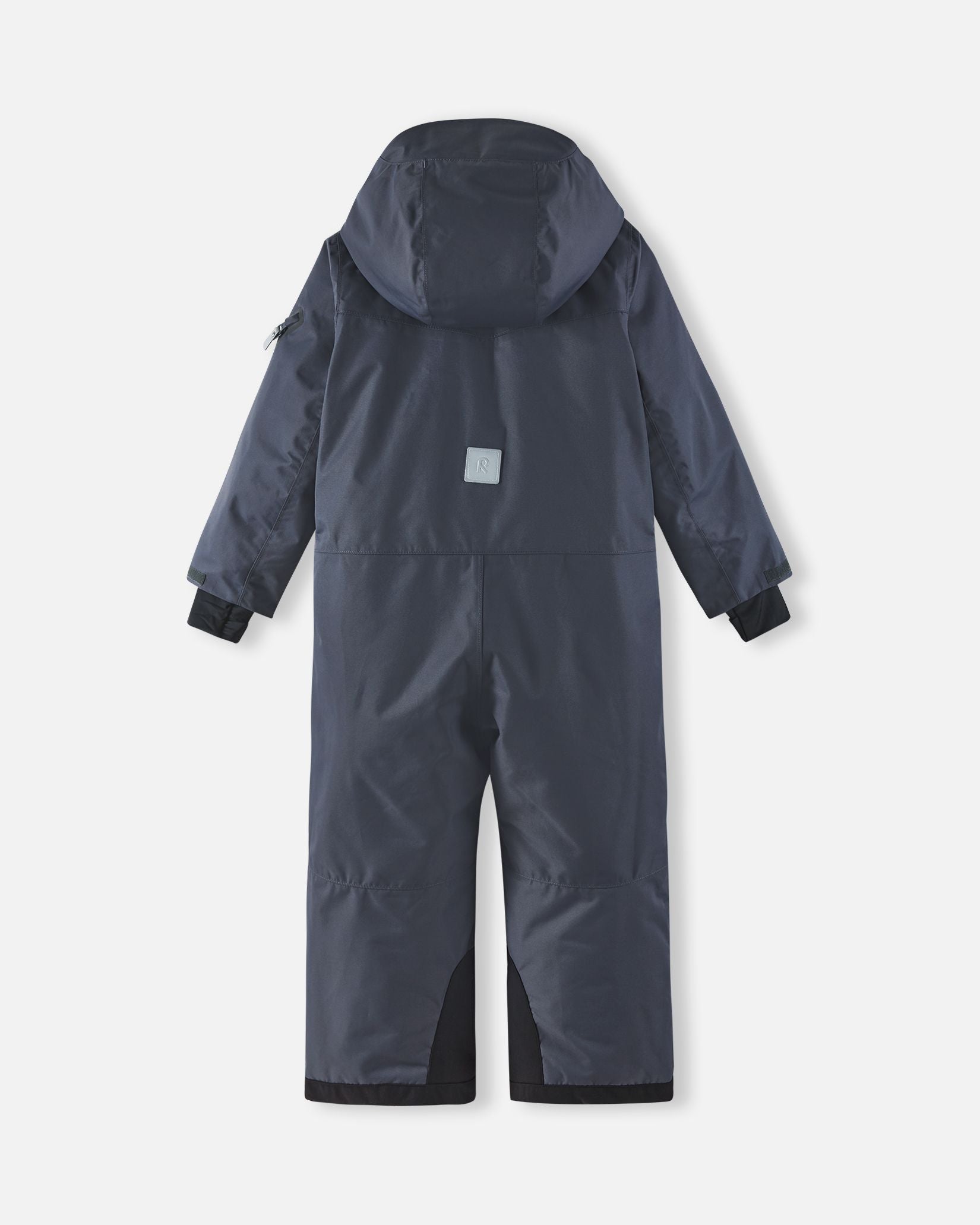Full back view of the Reima Reach kids' ski suit in soft black, highlighting the attached winter hood, a high-visibility reflective safety patch centered on the back, and heavy-duty black reinforced seat and ankle panels.
