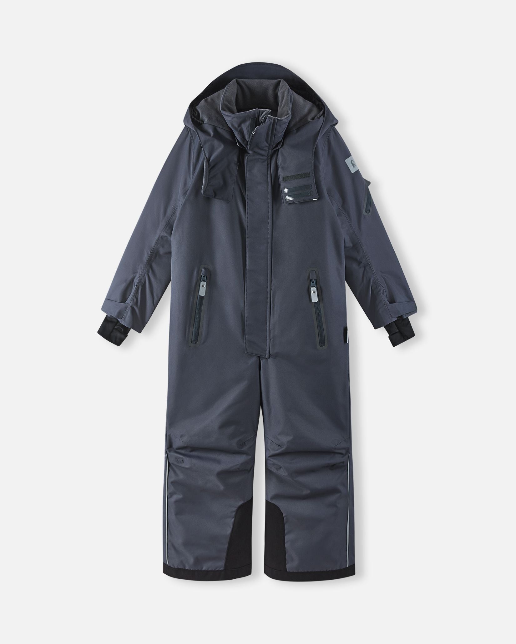 Full front view of the soft black Reima Reach kids' waterproof ski suit with the attached hood resting down, showcasing the protective high collar, dual zippered chest pockets, and highly durable black reinforced snow gaiters on the lower legs.