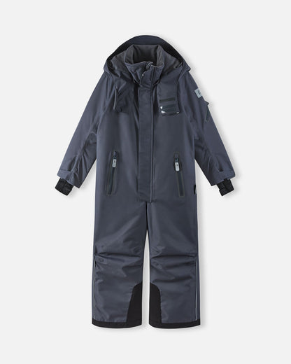 Full front view of the soft black Reima Reach kids' waterproof ski suit with the attached hood resting down, showcasing the protective high collar, dual zippered chest pockets, and highly durable black reinforced snow gaiters on the lower legs.