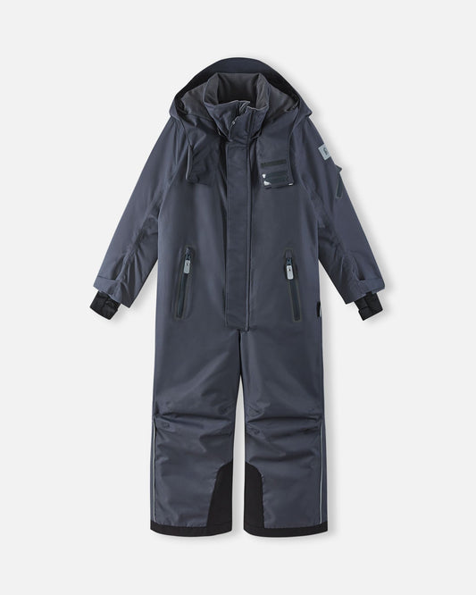 Full front view of the soft black Reima Reach kids' waterproof ski suit with the attached hood resting down, showcasing the protective high collar, dual zippered chest pockets, and highly durable black reinforced snow gaiters on the lower legs.