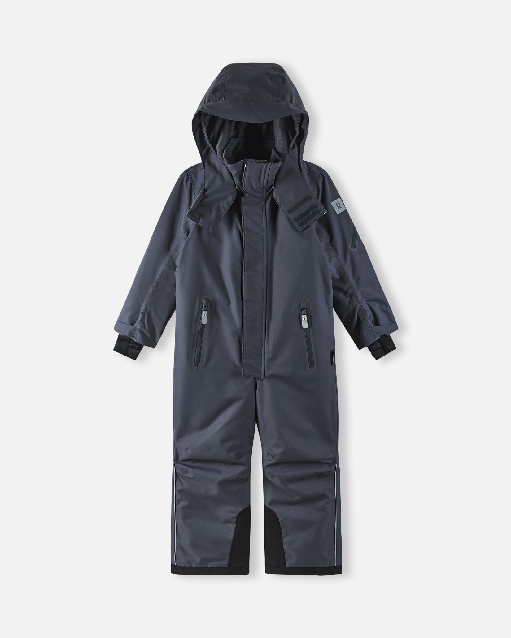 Full front view of the soft black Reima Reach kids' winter ski suit with the helmet-compatible hood pulled up, demonstrating the full-coverage weather protection and ergonomic design ideal for active winter sports.