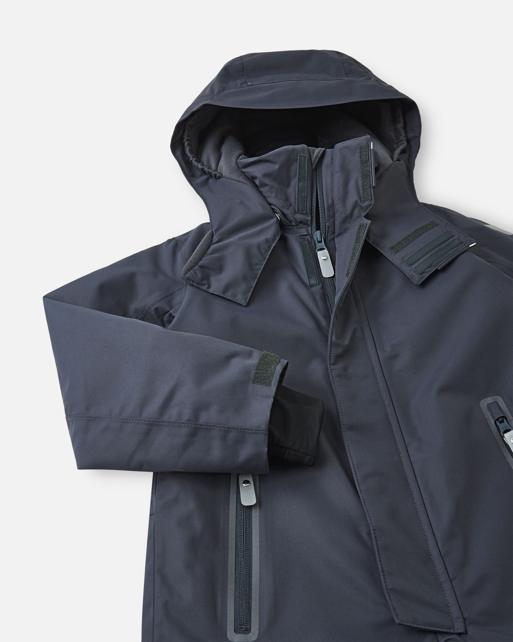 Detailed upper front view of the soft black Reima Reach kids' waterproof ski suit, highlighting the protective winter hood, high wind-blocking collar, and durable zippered chest pockets designed for active snow sports.