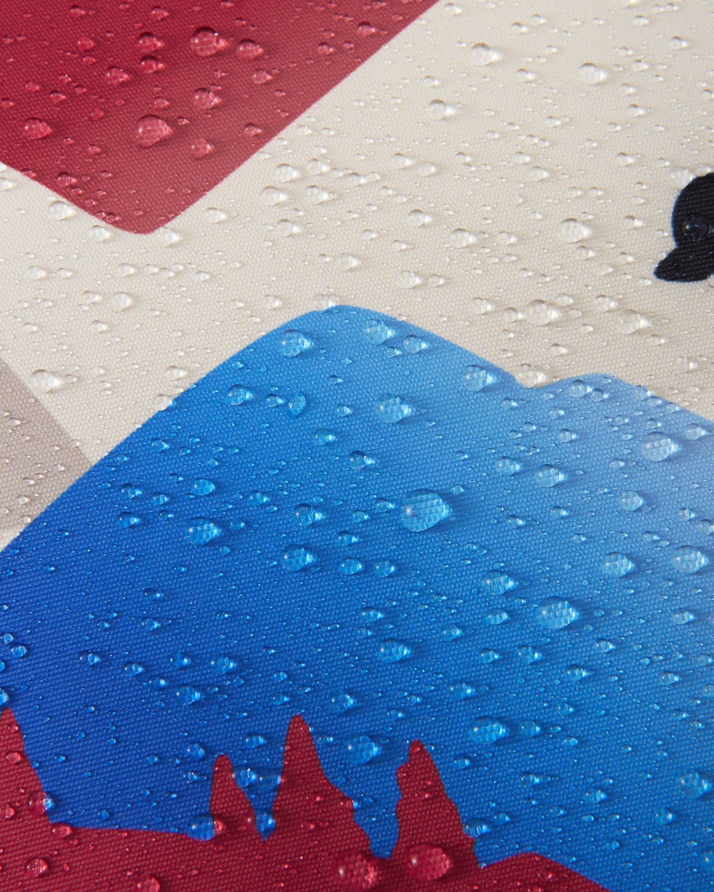 Macro close-up of water droplets tightly beading and resting on the blue, red, and cream Reimatec fabric of the Reach ski suit, clearly demonstrating the material's advanced waterproof and weather-resistant performance for winter conditions.
