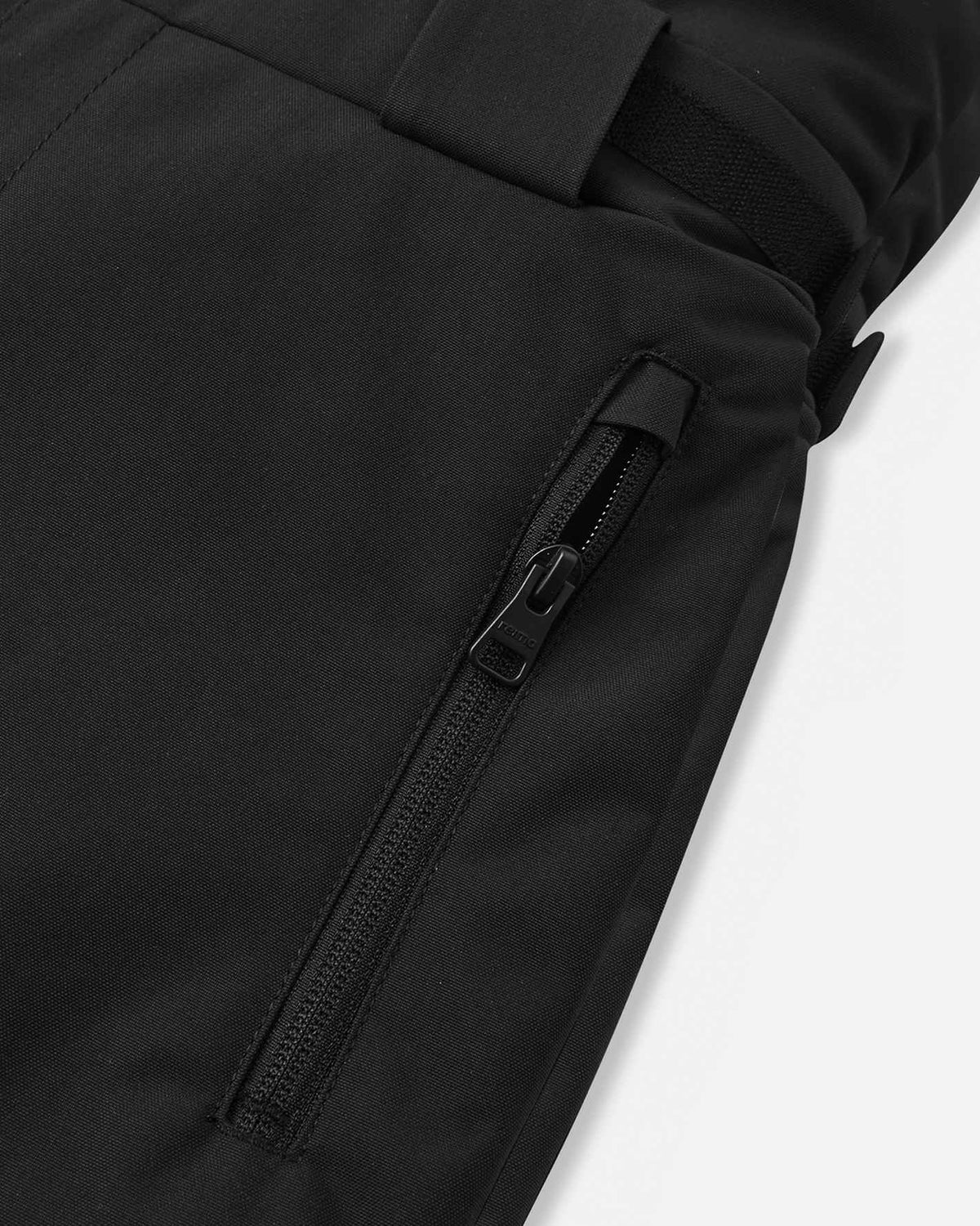 Detail view of the side waist and thigh on the black Reima Rehti kids' ski bib pants, showcasing the adjustable hook-and-loop waist tabs, ribbed side expansion panels for flexible movement, and a spacious cargo pocket.