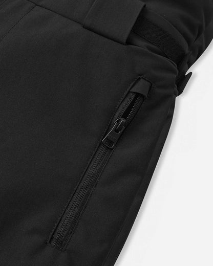 Detail view of the side waist and thigh on the black Reima Rehti kids' ski bib pants, showcasing the adjustable hook-and-loop waist tabs, ribbed side expansion panels for flexible movement, and a spacious cargo pocket.