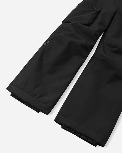 Detail view of the lower legs on the black Reima Rehti kids' ski bib pants, emphasizing the heavy-duty reinforced hems to protect against ski edges and internal elastic snow locks designed to secure over boots and block powder.
