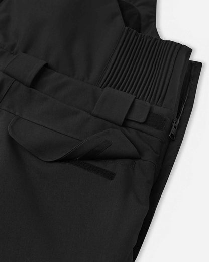 Close-up detail of a secure, low-profile zippered hip pocket on the black Reima Rehti kids' snow ski bibs, designed to keep small personal items safe and dry on the slopes.