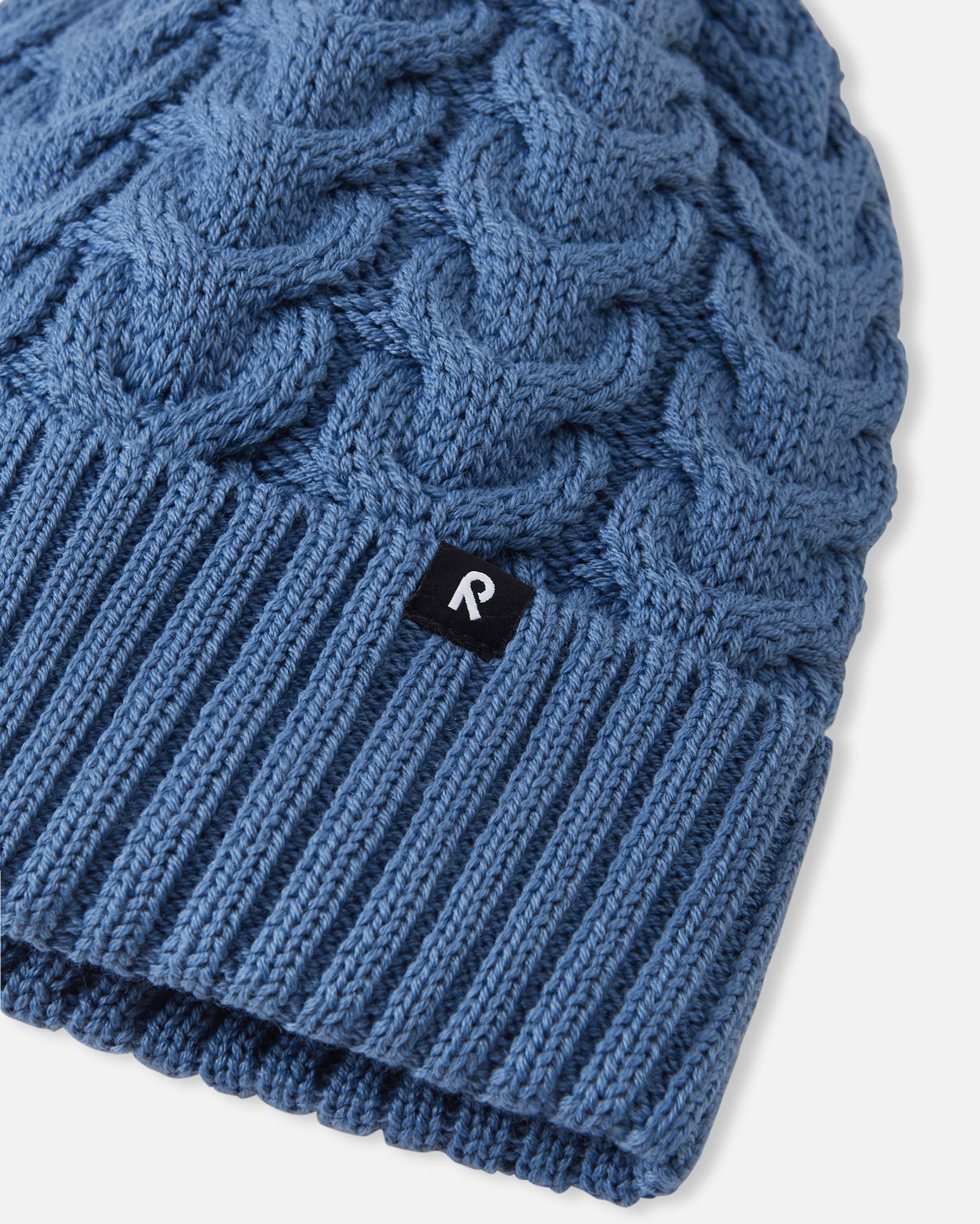 Close-up macro detail of the ribbed fold-up hem on the blue ocean Reima Routii kids' beanie, featuring the woven black Reima reflective label and the premium merino wool-mix yarn texture.