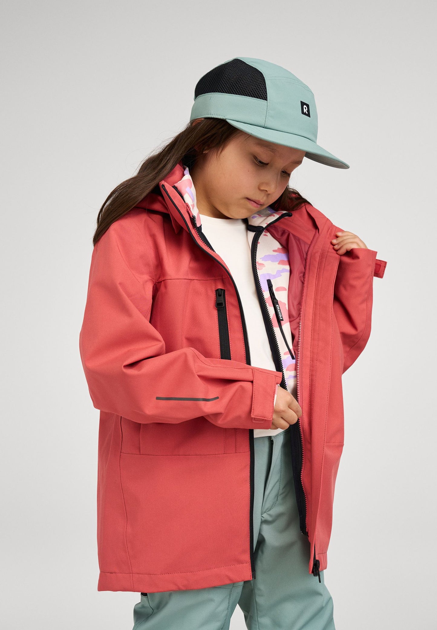 Lifestyle shot of a child zipping up the red clay Reima Saarela shell jacket over a patterned mid-layer, demonstrating the jacket's easy fit and comfortable layering capabilities for outdoor play.