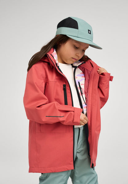Lifestyle shot of a child zipping up the red clay Reima Saarela shell jacket over a patterned mid-layer, demonstrating the jacket's easy fit and comfortable layering capabilities for outdoor play.