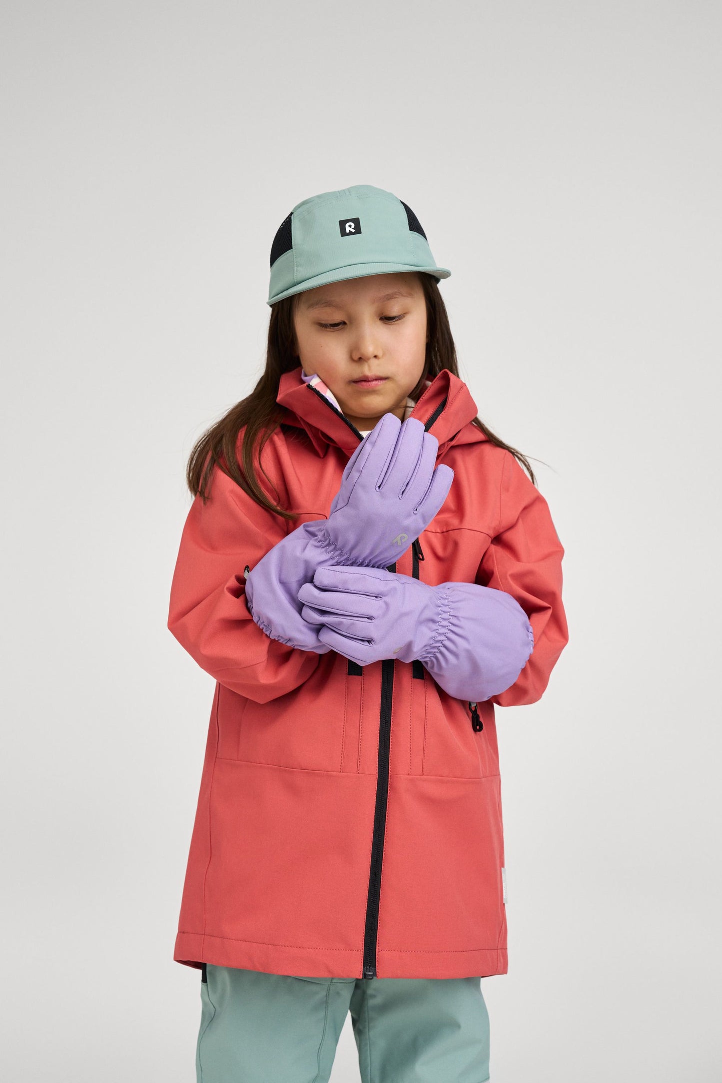 Lifestyle shot of a child wearing the red clay Reima Saarela shell jacket paired with purple Reima winter gloves and a stone green cap, showcasing a complete, weather-ready outdoor outfit.