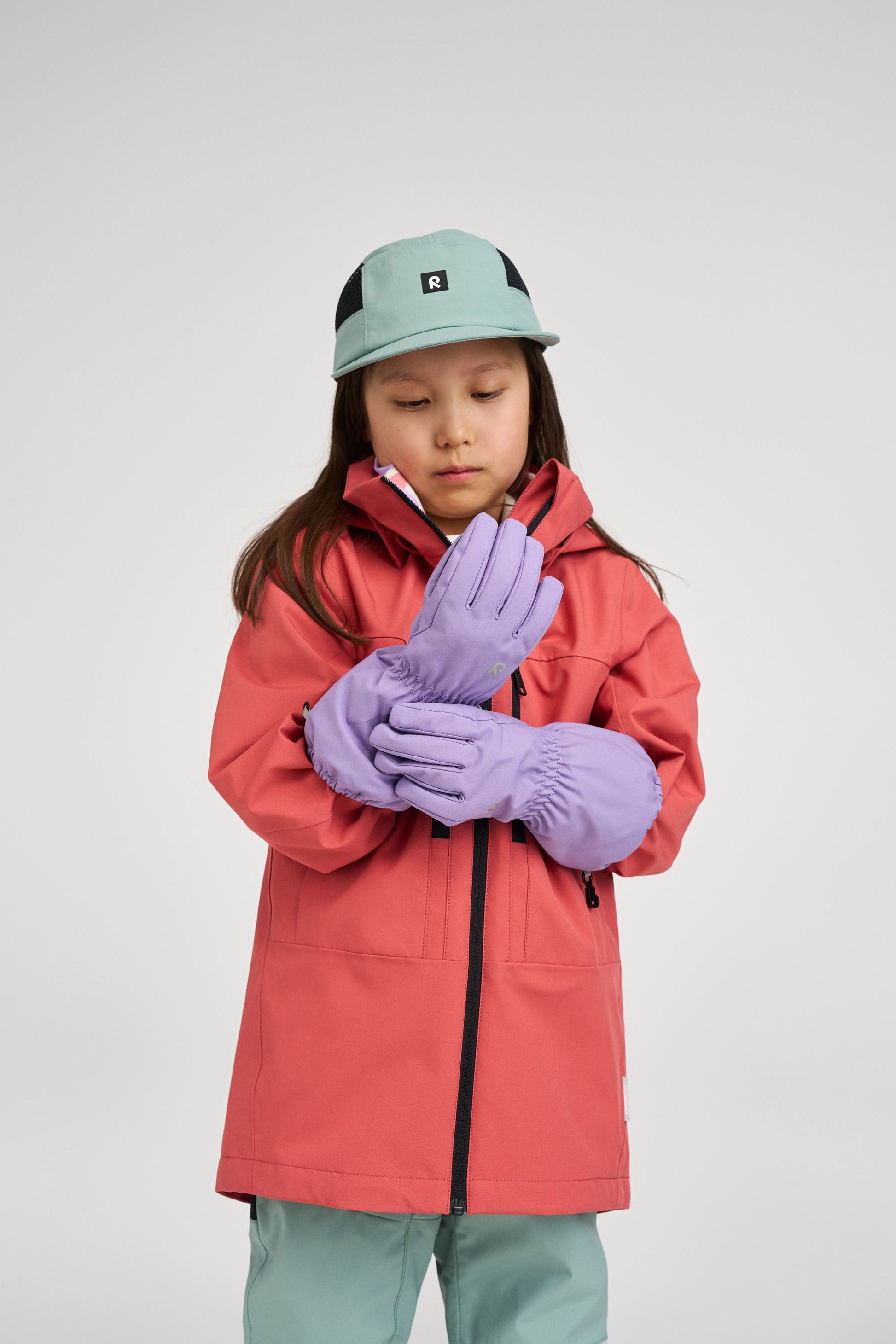 Lifestyle shot of a child wearing the red clay Reima Saarela shell jacket paired with purple Reima winter gloves and a stone green cap, showcasing a complete, weather-ready outdoor outfit.