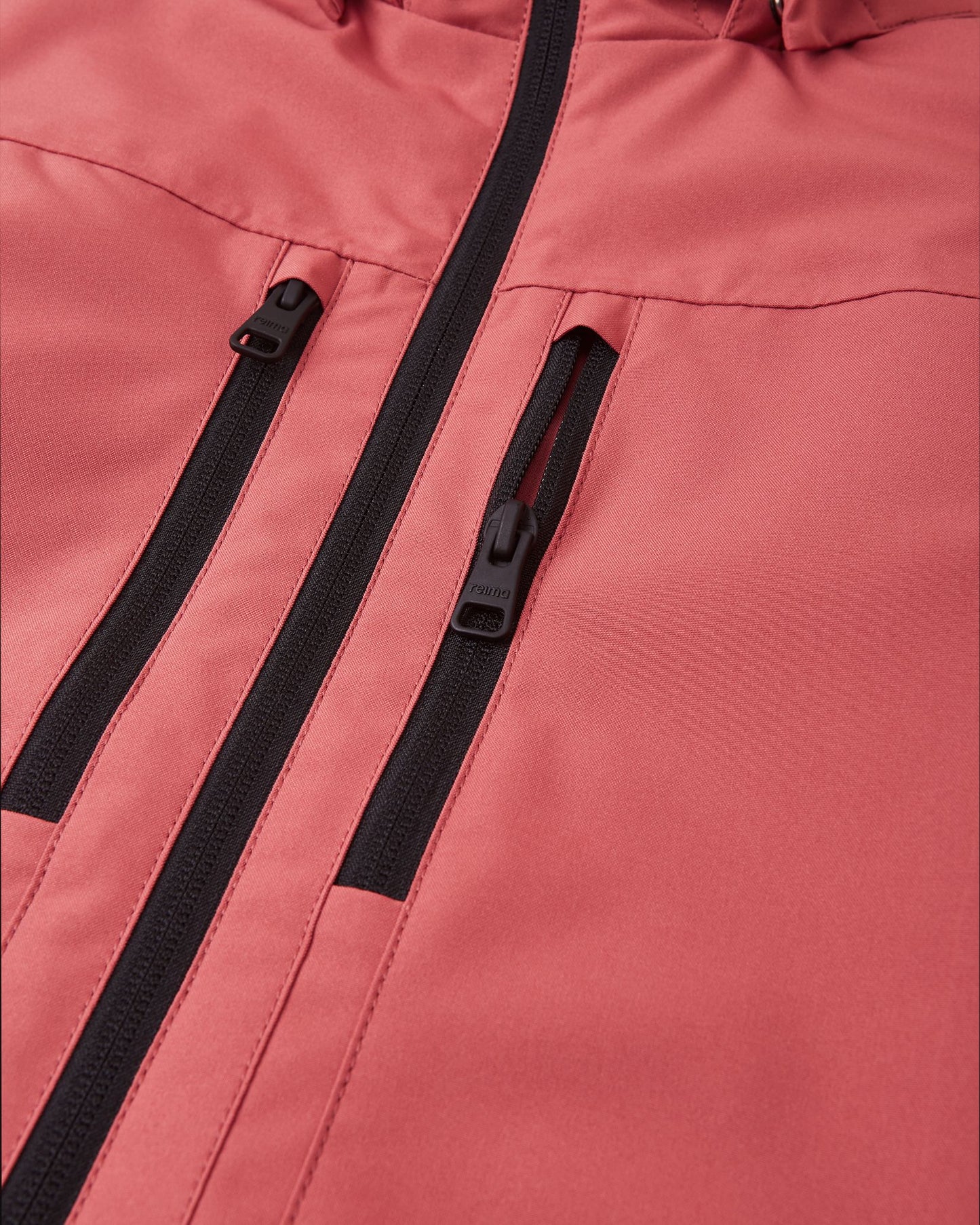 Close-up detail of the Reima Saarela kids' red clay shell jacket, highlighting the two secure zippered chest pockets with durable black hardware set against the waterproof fabric.
