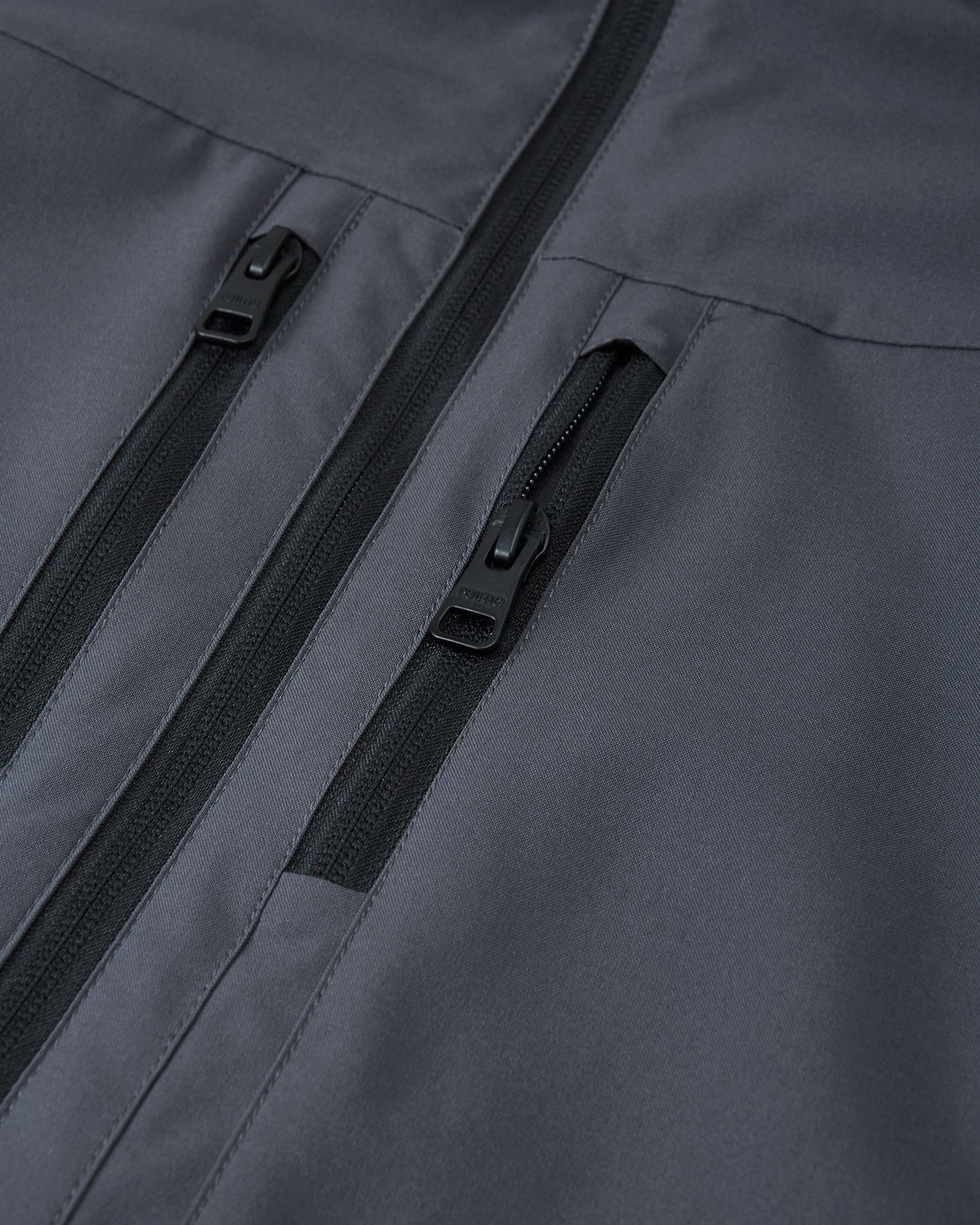 Close-up of the two secure, vertical zippered chest pockets on the Reima Saarela waterproof jacket in soft black.