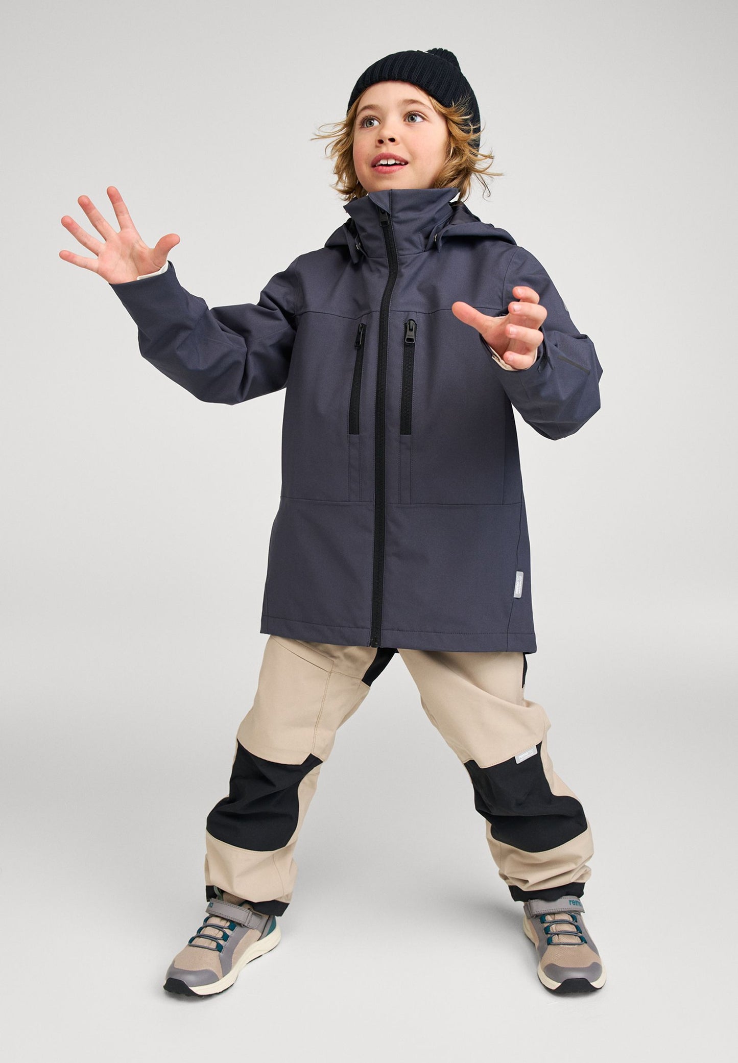 A child posing in the Reima Saarela kids waterproof jacket in soft black, paired with outdoor pants and a beanie for fall weather adventures.