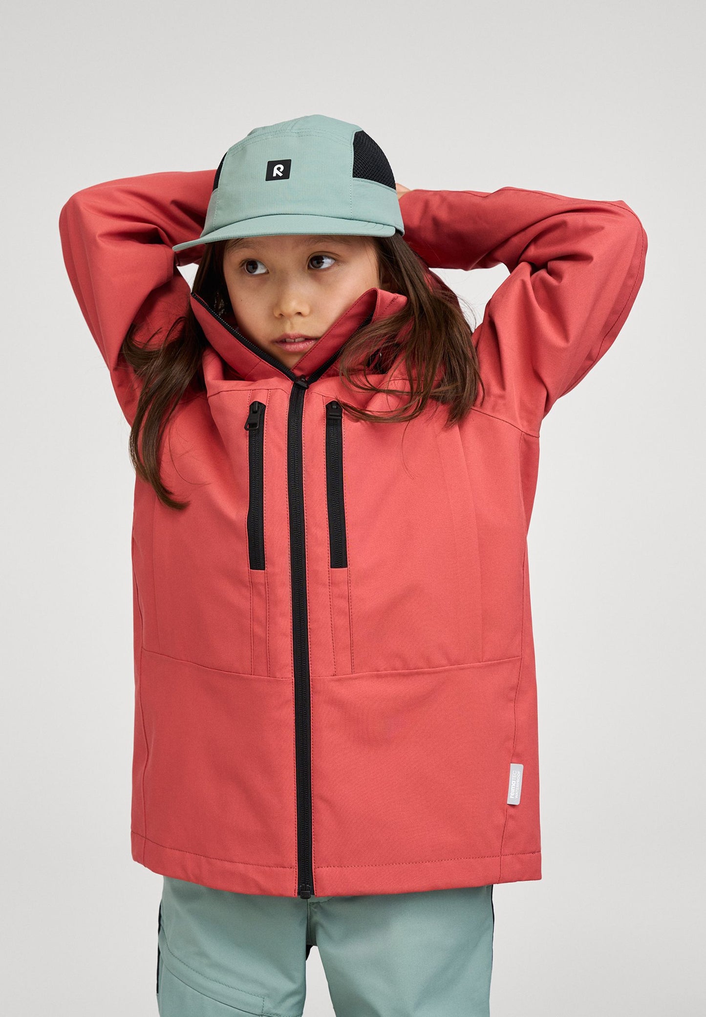 Lifestyle shot of a child wearing the red clay Reima Saarela kids' waterproof shell jacket over a stone green cap, holding the high collar up to demonstrate its protective, wind-blocking fit.