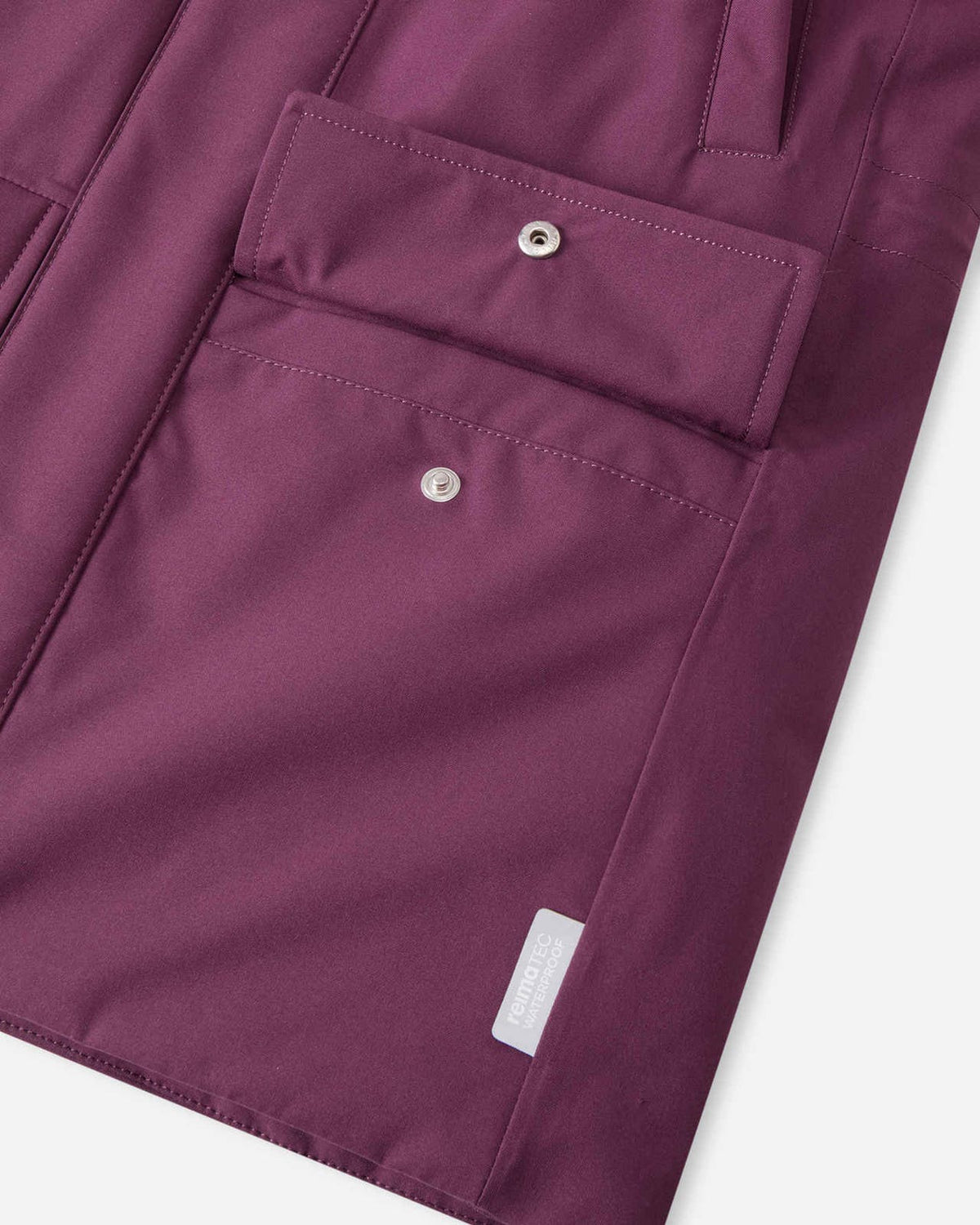 Macro detail of the front cargo pocket on the deep purple Reima Serkkula kids' parka, displaying the silver snap button closure and woven Reimatec waterproof tag.
