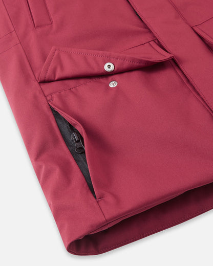 A close-up detail of the lower front pocket construction on the maroon Reima Serkkula kids' down parka, displaying a snap-button flap pocket paired with a hidden side zippered pocket for secure storage.