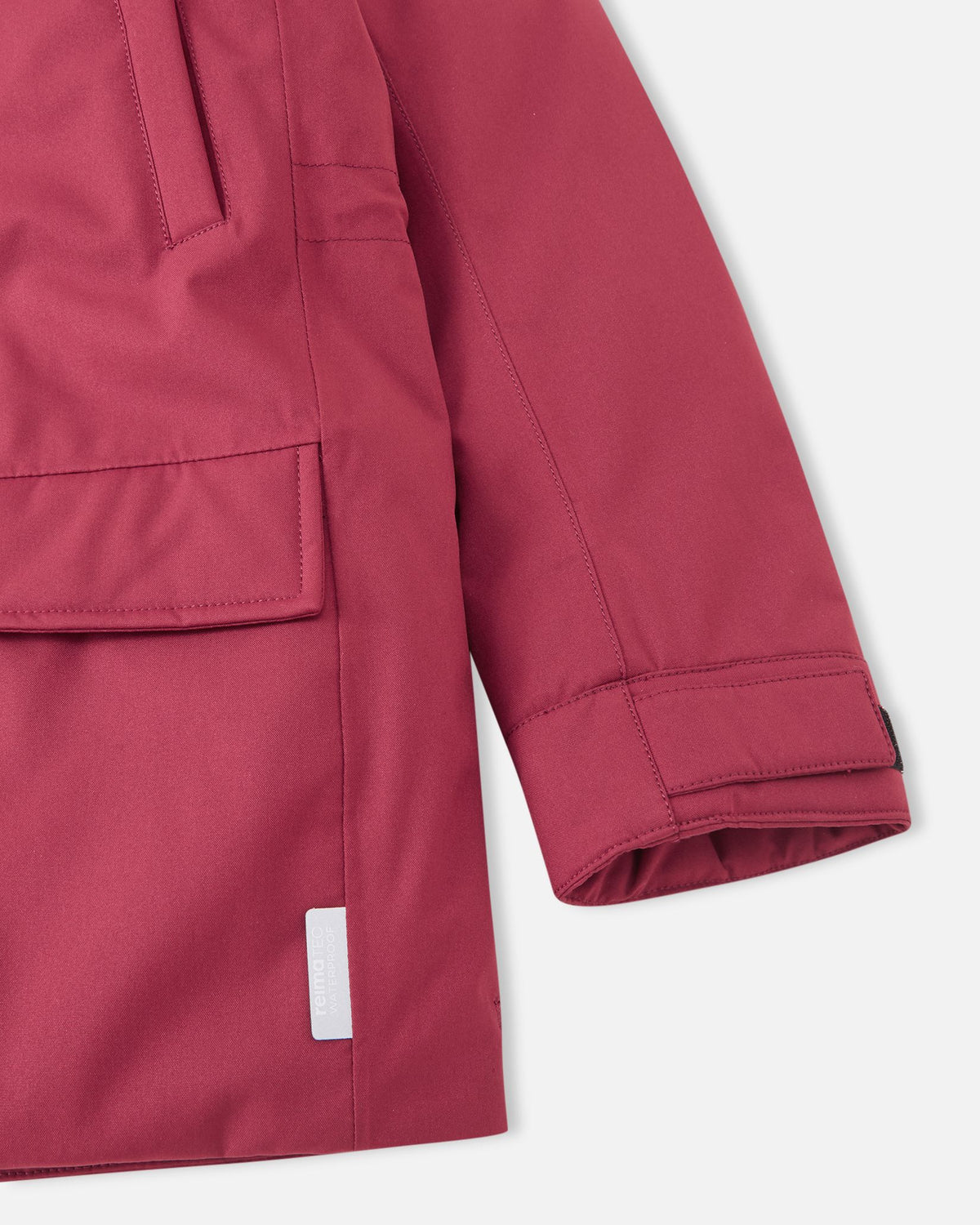 A detail close-up showing the bottom hem and side pocket flap of the maroon Reima Serkkula kids' parka, including a visible white reflective safety tag near the hem.