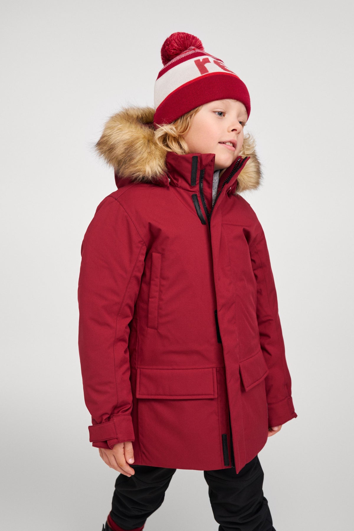 A three-quarter side view of a young child model wearing the maroon Reima Serkkula kids' down parka jacket and a Reima knit hat, seen on a light grey studio background.