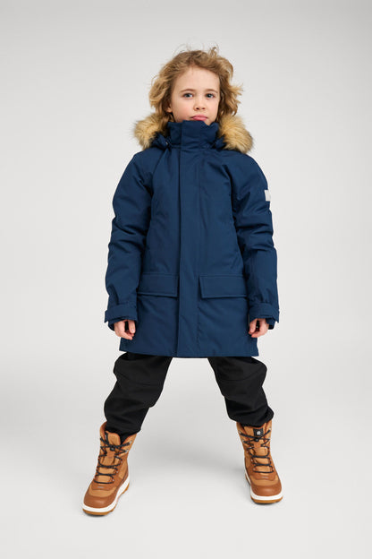 Full-length view of a child model wearing the navy blue Reima Serkkula kids' down parka jacket, black pants, and winter boots against a studio background.