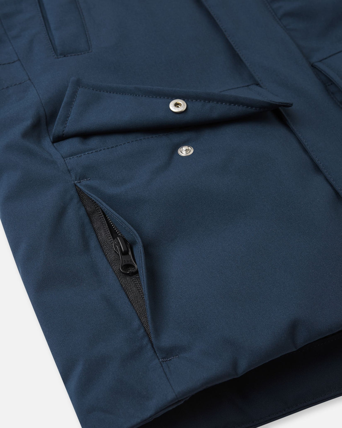 A close-up detail of the lower front pocket construction on the navy blue Reima Serkkula kids' down parka, showing a snap-button flap pocket paired with a hidden side pocket with a zipper for secure storage.
