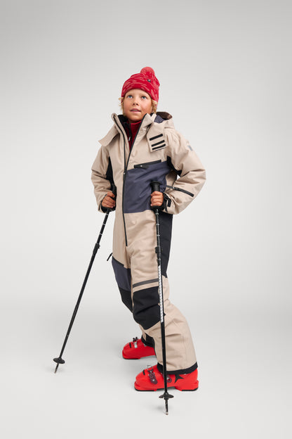 A young boy wearing the Reima Skaulo kids ski suit in earthy beige, standing with ski poles and red ski boots, ready for winter alpine skiing.