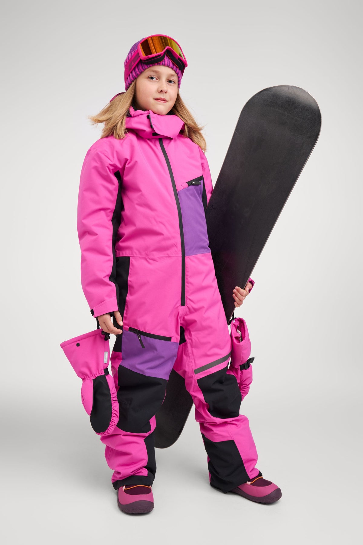 A young girl ready for winter sports wearing the Reima Skaulo girls ski suit in fuchsia, holding a snowboard and matching winter mittens attached to the suit.