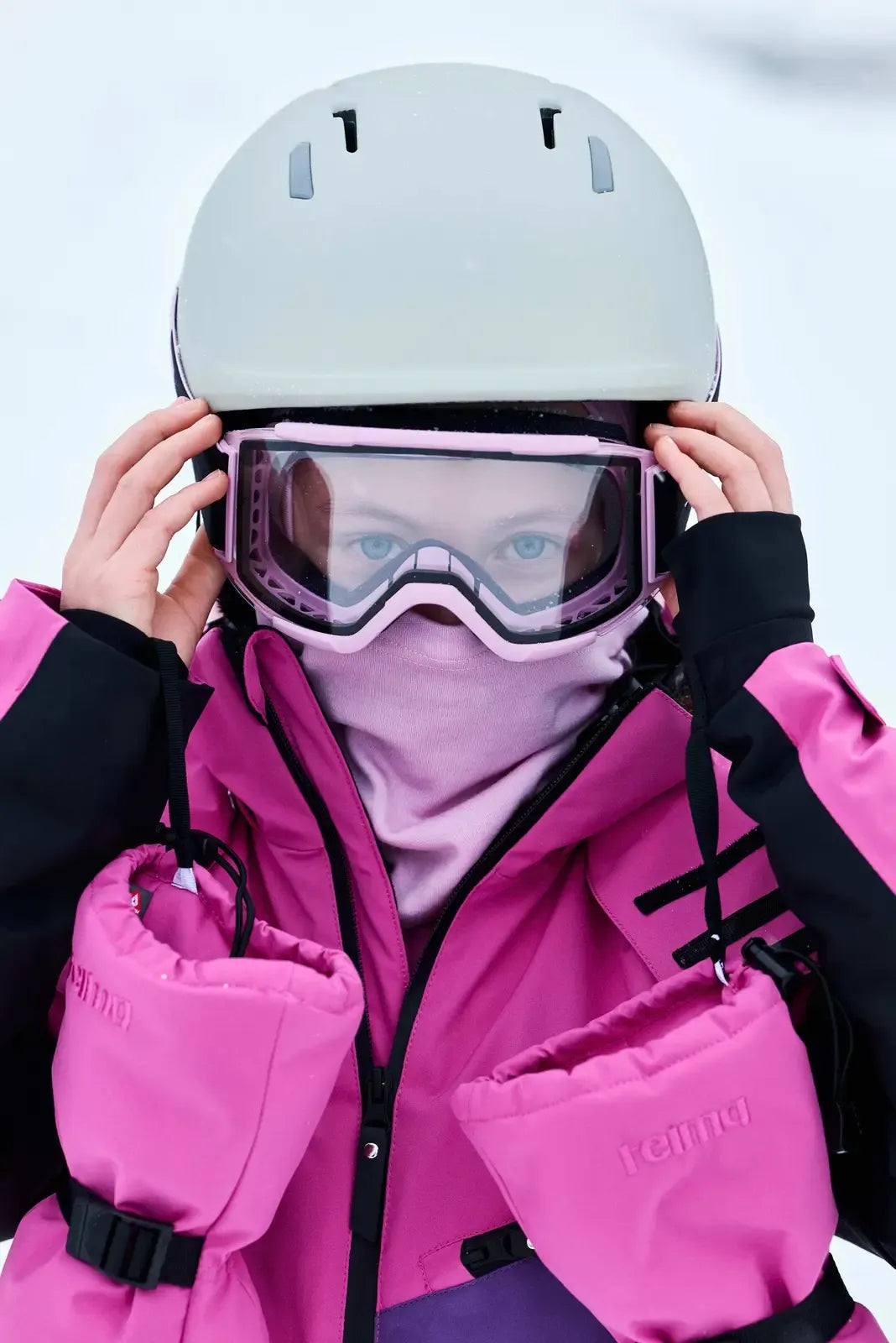 A girl adjusting her ski goggles and helmet while wearing the fuchsia Reima Skaulo kids' ski suit and matching Reima Vanukko mittens.
