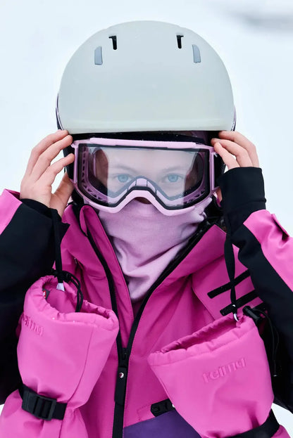 A girl adjusting her ski goggles and helmet while wearing the fuchsia Reima Skaulo kids' ski suit and matching Reima Vanukko mittens.