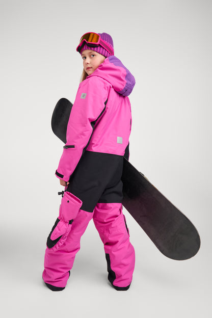 Back and side angle of a girl modeling the fuchsia Reima Skaulo girls ski suit, holding a snowboard and demonstrating the durable black reinforcement panels on the seat and legs.