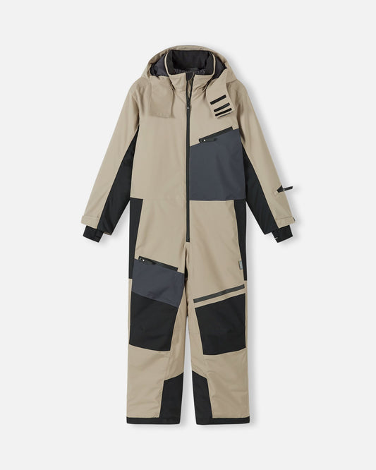 Front view of the Reima Skaulo kids ski suit in earthy beige, featuring a waterproof exterior, black color-blocked reinforcement panels on the knees and arms, and a dark grey zippered chest pocket.