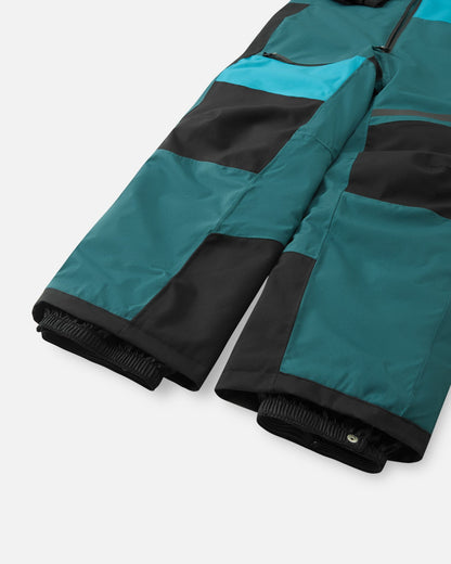Detail shot of the lower legs on the Reima Skaulo kids' ski suit, displaying the rugged black reinforced knee and shin panels, alongside the inner elastic snow gaiters with snap closures designed to stretch over ski boots and keep snow out.