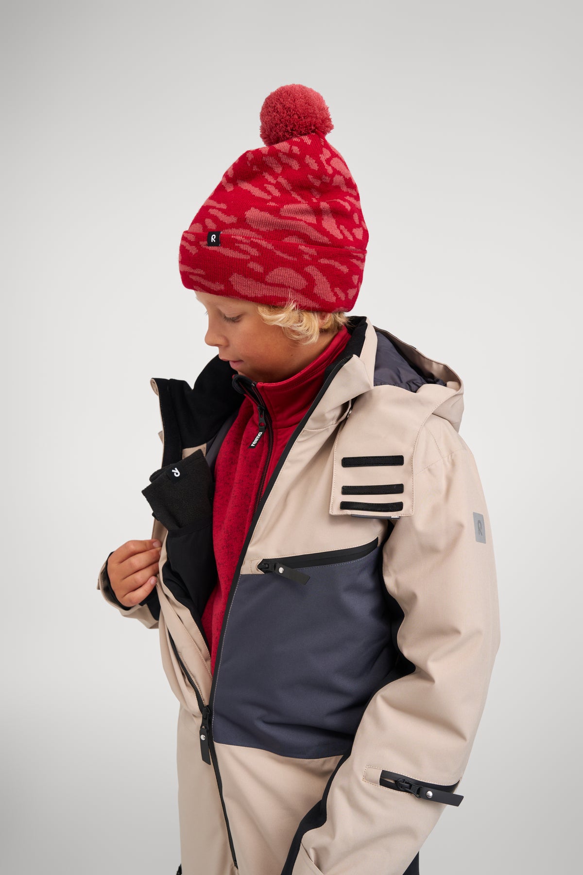 Side view of a boy opening the front zip of the earthy beige Reima Skaulo kids ski suit, revealing a warm red mid-layer and demonstrating the comfortable, ergonomic fit.
