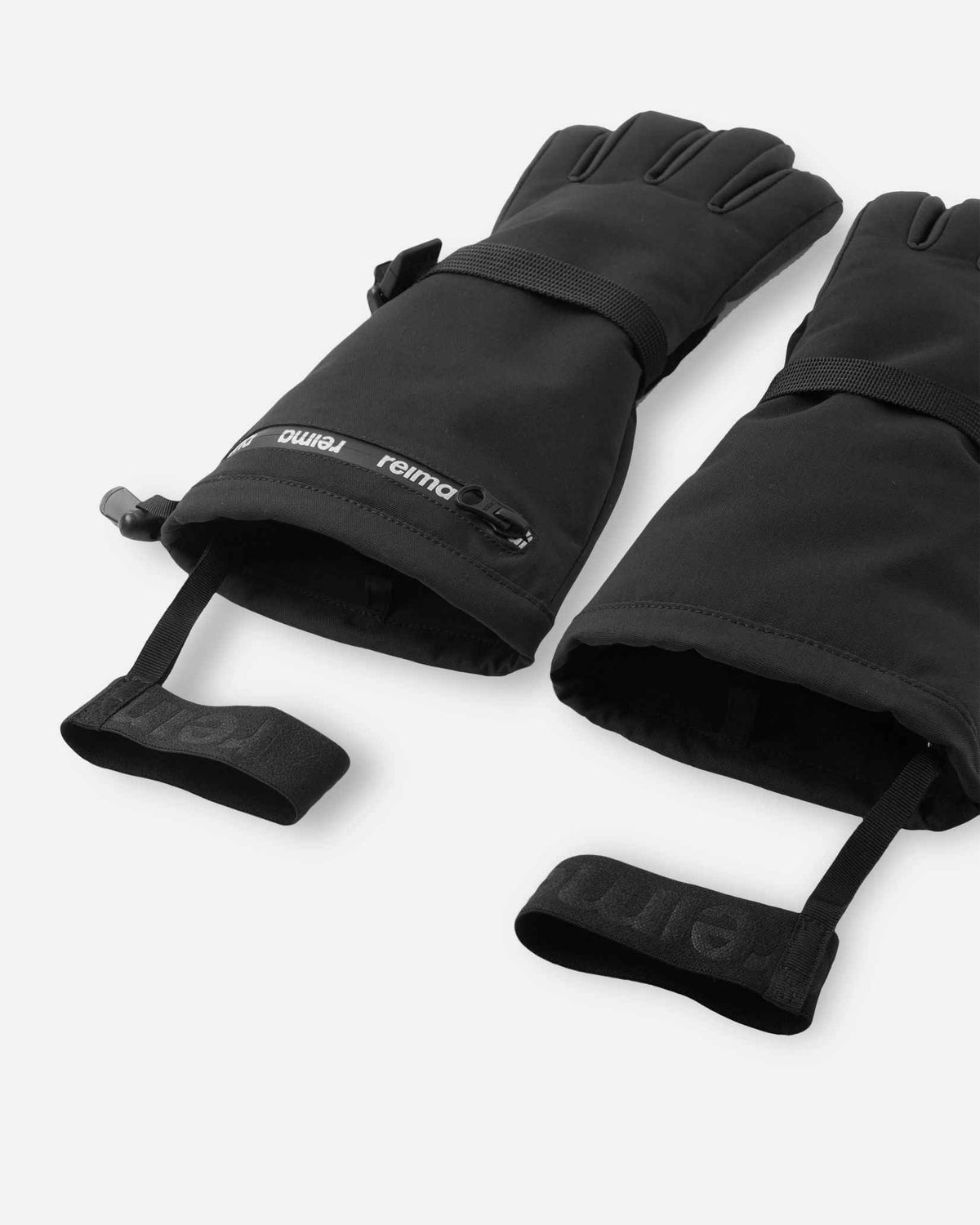 Angled flat lay view of the black Reima Skimba children's ski gloves, emphasizing the adjustable wrist straps, water-resistant zippered pockets, and attached elastic safety wrist leashes.