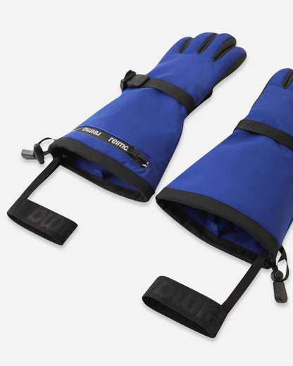 Angled flat lay view of the twilight blue Reima Skimba children's ski gloves, emphasizing the adjustable wrist straps, water-resistant zippered pockets, and attached elastic safety wrist leashes.