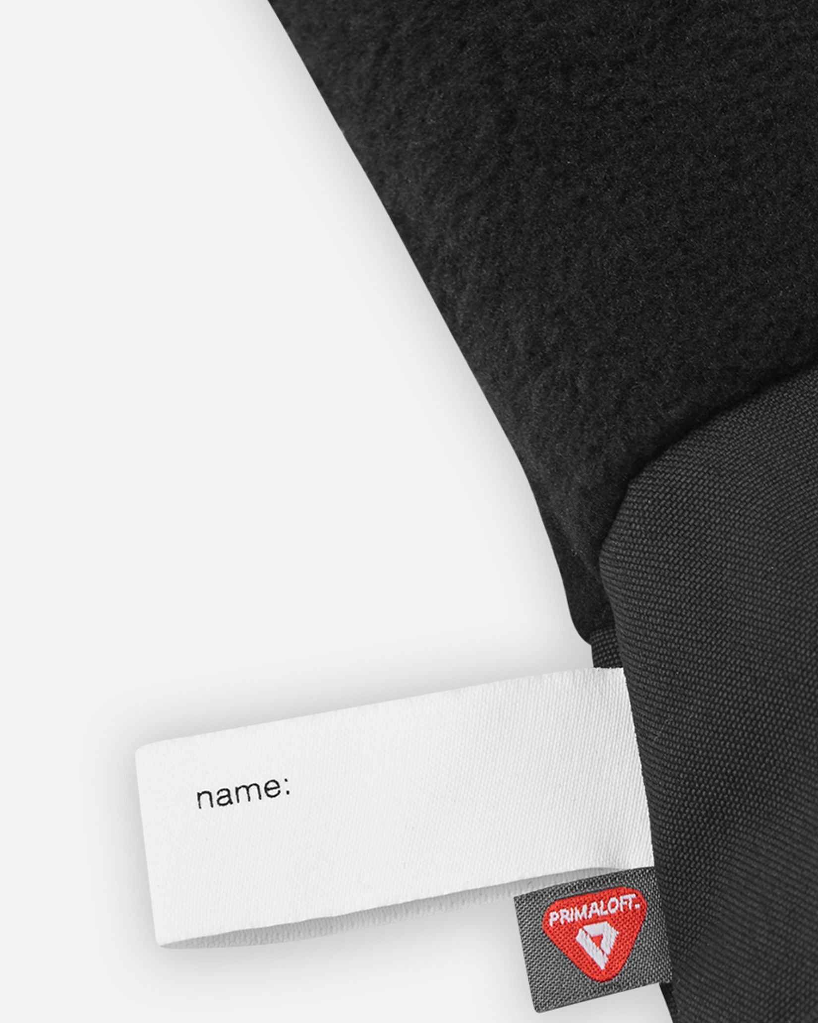 Detail of the inner cuff of the black Reima Skimba kids' ski gloves, showing the soft thermal fleece lining, a customizable blank name tag, and the premium PrimaLoft insulation label indicating high-performance winter warmth.