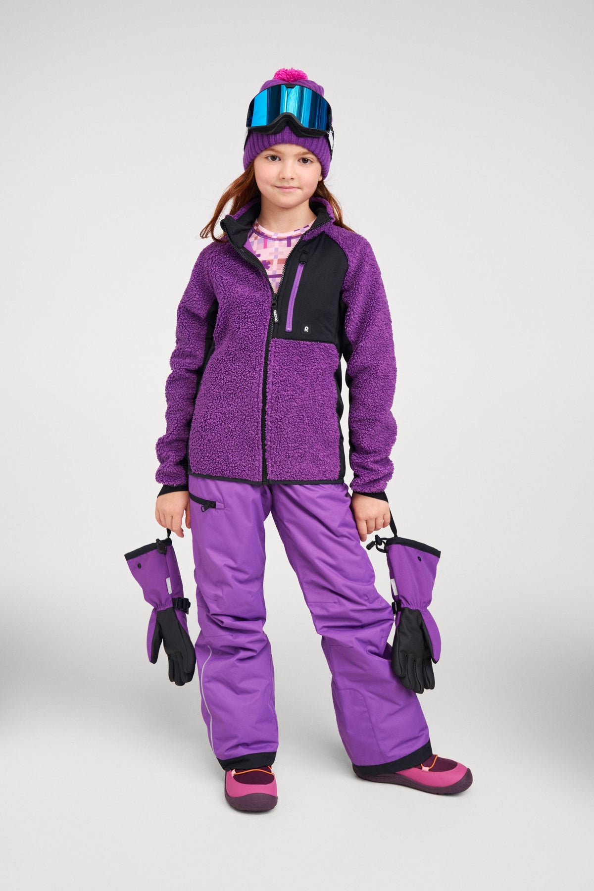 Lifestyle portrait of a young skier in a full purple winter outfit and ski goggles, demonstrating the elastic safety wrist leashes on her plum purple Reima Skimba kids' ski gloves as they hang securely from her wrists.