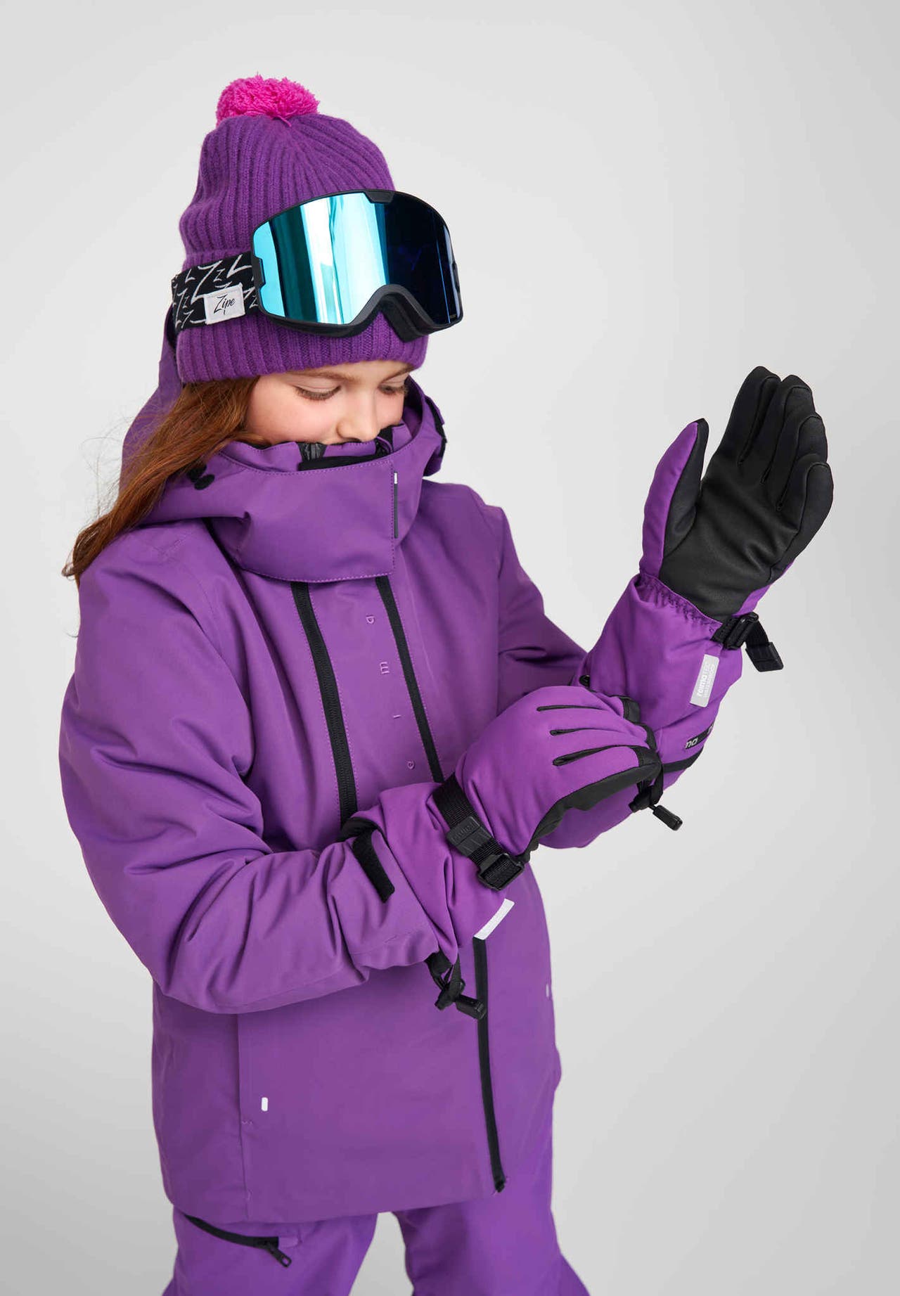 Lifestyle portrait of a young girl wearing a purple winter jacket, ribbed beanie, and mirrored ski goggles, actively adjusting her plum purple Reima Skimba kids' ski gloves. The image highlights the gloves' secure adjustable wrist straps and flexible fingered design engineered for optimal grip and dexterity on the slopes.