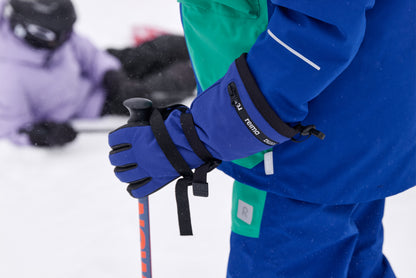 Close-up lifestyle action shot of a young skier gripping a ski pole while wearing twilight blue Reima Skimba kids' ski gloves. The image demonstrates how the extra-long zippered gauntlet cuffs and adjustable wrist straps layer perfectly over a winter jacket sleeve to completely block out snow during alpine skiing.